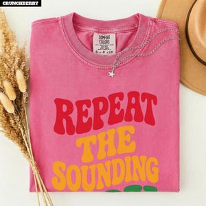 May include: A pink short-sleeve t-shirt with a crew neckline and a Comfort Colors label. The front features the text REPEAT THE SOUNDING in large, wavy red and yellow letters. A silver-toned chain necklace with a small star charm rests on the shirt.