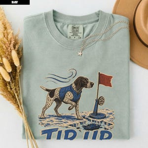 May include: A sage green crewneck t-shirt featuring a graphic of a spotted dog wearing a blue vest standing on ice near a red flag and a fishing hole. The text "TIP UP" is printed below the graphic. The shirt has a Comfort Colors brand label.