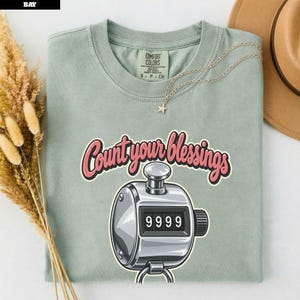 Peut inclure: Un t-shirt à manches courtes vert sauge avec un graphique représentant un compteur manuel argenté affichant le nombre 9999. Au-dessus du compteur, le texte Count your blessings est imprimé en police cursive rose avec un contour foncé.