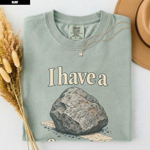 Op de afbeelding: Een saliegroen T-shirt met ronde hals en de tekst I have a boven een grafische illustratie van een grote grijze rots. Het shirt heeft een Comfort Colors-label bij de halslijn en wordt getoond met een gouden sterrenketting en een beige hoed.
