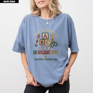 May include: A blue jean coloured t-shirt featuring a graphic print with the text "TEACH THE FIRST STEPS ON HOLIDAY DUTY" and "PRESCHOOL TEACHER CREW". The design includes illustrations of a crayon, alphabet blocks, scissors, and small footprints.
