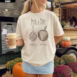 以下が含まれることがあります： Pick A Sideというテキストがプリントされたアイボリーの半袖Tシャツ。テキストの下には、No. I stuffingとラベル付けされた詰め物入りのスプーンと、No. II dressingとラベル付けされたドレッシング入りのスキレットの2つのイラストが描かれています。