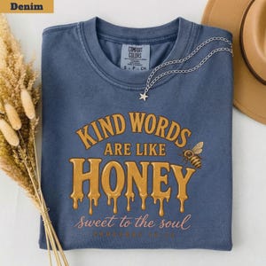 以下が含まれることがあります： 「KIND WORDS ARE LIKE HONEY sweet to the soul Proverbs 16:24」という金と白の文字がプリントされたデニムブルーのクルーネックTシャツ。デザインには小さなハチのイラストが含まれており、生地の上には銀色の星のペンダントネックレスが置かれています。