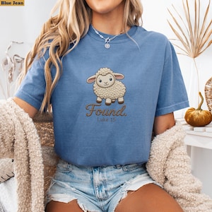 Puede incluir: Una camiseta azul con el gráfico de una oveja de color crema. Debajo de la oveja, el texto dice "Found." y "Luke 15" en una fuente cursiva marrón. La camiseta tiene cuello redondo y mangas cortas, ofreciendo un ajuste casual y cómodo.
