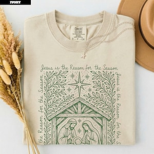 May include: An ivory t-shirt featuring a dark green graphic print of a nativity scene inside a stable, topped with a star and surrounded by leafy branches. The text "Jesus is the Reason for the Season" is printed in a decorative font around the design.