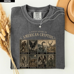 Puede incluir: Una camiseta gris carbón con un gráfico titulado A Field Guide to American Cryptids. El estampado muestra ocho ilustraciones enmarcadas de criaturas legendarias, incluyendo Sasquatch, Mothman, Champ, Jackalope, Wendigo, Skinwalker, Jersey Devil y Flatwoods Monster.
