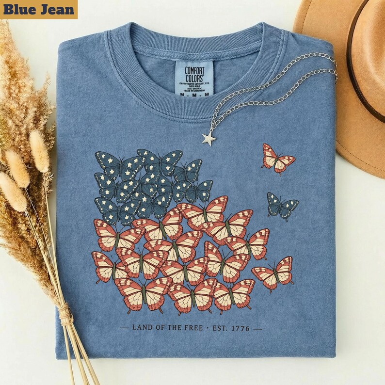 以下が含まれることがあります： 青いジーンズ色のTシャツ。赤、白、青の蝶のデザインで、アメリカの国旗を模倣しています。Tシャツはクルーネックで半袖です。「LAND OF THE FREE - EST. 1776」の文字が下部にプリントされています。