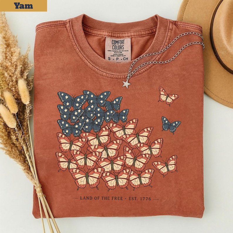 以下が含まれることがあります： アメリカ国旗の形をした蝶のデザインが特徴の、錆色のTシャツ。星には青と白の蝶、縞には赤と白の蝶が使われています。「LAND OF THE FREE - EST. 1776」の文字が下部にプリントされています。