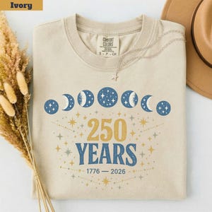 Op de afbeelding: Ivoor kleurig T-shirt met een ronde hals. Het shirt heeft een hemels ontwerp met maanfasen en sterren, samen met de tekst "250 YEARS" en de data "1776 - 2026". Een zilveren ketting met een ster hanger ligt op het shirt.