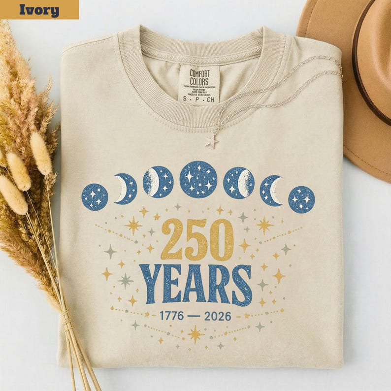 Op de afbeelding: Ivoor kleurig T-shirt met een ronde hals. Het shirt heeft een hemels ontwerp met maanfasen en sterren, samen met de tekst "250 YEARS" en de data "1776 - 2026". Een zilveren ketting met een ster hanger ligt op het shirt.