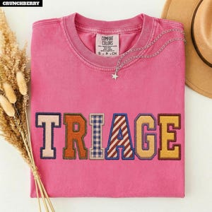 May include: A pink short-sleeve t-shirt featuring the word TRIAGE in large, multi-patterned embroidered letters across the chest. The letters include solid colors, a blue and white gingham check, and red and white diagonal stripes. A silver star necklace rests on the collar.