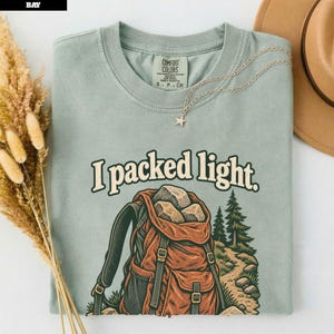 May include: A sage green short-sleeve t-shirt featuring a graphic of an orange backpack filled with rocks, a forest path, and pine trees. The text "I packed light." is printed above the backpack. The shirt has a Comfort Colors brand label.