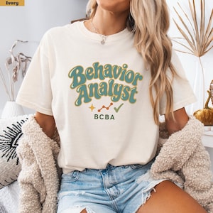 Pode incluir: Uma t-shirt de manga curta na cor marfim com o texto "Behavior Analyst" numa fonte retro em tom verde-azulado com contorno dourado. Abaixo do texto, há uma pequena estrela dourada, um gráfico de linhas dourado, um visto verde e "BCBA".