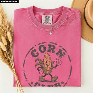 Könnte beinhalten: Ein rosafarbenes Kurzarm-T-Shirt mit einer verwaschenen kreisförmigen Grafik, dem Text CORN und einer Cartoon-Maiskolben-Figur, die den Daumen hochhält. Das Shirt hat ein Comfort Colors-Etikett am Kragen und eine silberne Stern-Halskette.