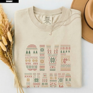 May include: An ivory short-sleeve t-shirt featuring a graphic print with the word GIVE in large letters filled with red, green, and gold holiday-themed patterns, including trees, snowflakes, and hearts, along with the full alphabet in small letters.