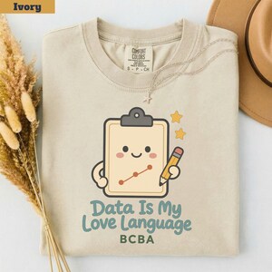 Op de afbeelding: Een ivoorkleurig t-shirt met een afbeelding van een lachend klembord dat een potlood vasthoudt, met een lijngrafiek op het oppervlak. Onder de afbeelding staat de tekst "Data Is My Love Language" in blauw en "BCBA" in groen. Het t-shirt heeft een ronde hals.