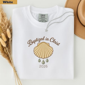 May include: A white t-shirt featuring an embroidered design of a tan scallop shell with the text "Baptized in Christ" arched above it, three small green water droplets below the shell, and the year "2026" printed at the bottom center.