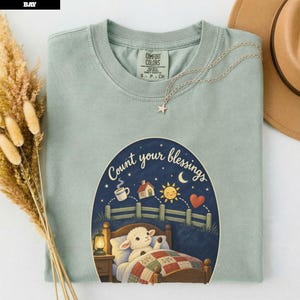 Peut inclure: Un t-shirt vert sauge avec une illustration représentant un agneau au lit sous un ciel étoilé et le texte Count your blessings. Le motif inclut une maison, un soleil, un cœur et une lanterne. Un collier étoile argenté repose sur le col.