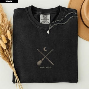 May include: A black crewneck t-shirt featuring a graphic of two crossed broomsticks and a crescent moon. Below the graphic, the text reads "basic witch". The shirt has a Comfort Colors label inside the collar and a silver star necklace.