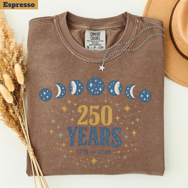 Op de afbeelding: Espresso-kleurig T-shirt met een hemels ontwerp. Het toont maanfasen en sterren, met de tekst "250 YEARS" en de jaren 1776-2026. Een zilveren ketting met een sterhanger ligt op het T-shirt.