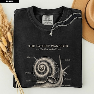 Könnte beinhalten: Ein schwarzes T-Shirt mit Rundhalsausschnitt und einer weißen anatomischen Zeichnung einer Schnecke. Der Text lautet THE PATIENT WANDERER und Cochlea umbralis, mit Beschriftungen für spire, aperture, mantle, tentacle und operculum. Comfort Colors Etikett.
