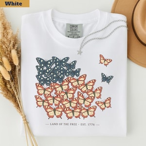 以下が含まれることがあります： アメリカ国旗を模した蝶のデザインが施された白いTシャツ。星を表す青と白の蝶、縞を表す赤とタンの蝶を使用。デザインの下には「LAND OF THE FREE &bull; EST. 1776」の文字がプリントされています。