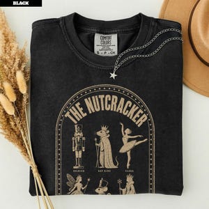 Peut inclure: Un t-shirt noir à col rond avec une impression graphique beige représentant les personnages de Casse-Noisette, dont un soldat, le roi des rats et une ballerine. Le texte indique The Nutcracker, Soldier, Rat King et Clara. Étiquette Comfort Colors.