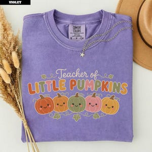 May include: A violet short-sleeve t-shirt with a graphic print that reads Teacher of LITTLE PUMPKINS in multi-coloured lettering. Below the text are five illustrated pumpkins with smiling faces in orange, yellow, green, and pink colours.