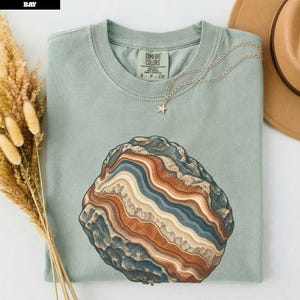 Pode incluir: Uma t-shirt verde sálvia com um gráfico de uma pedra de ágata com faixas em tons de azul, creme e laranja ferrugem. A t-shirt tem gola redonda e uma etiqueta Comfort Colors. Um delicado colar de corrente prateada com uma pequena estrela repousa sobre o tecido.