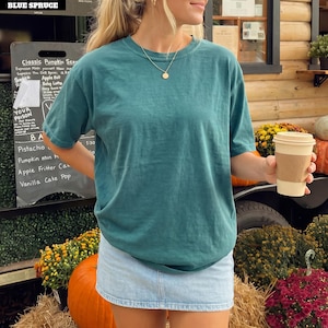 May include: A solid teal short-sleeve crewneck t-shirt paired with a light blue denim mini skirt. The t-shirt features a relaxed fit and a simple round neckline. A gold pendant necklace with a circular charm rests against the fabric of the shirt.