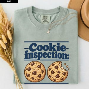 Op de afbeelding: Een saliegroen T-shirt met ronde hals en de donkerblauwe tekst Cookie inspection: boven twee chocoladekoekjes, waarvan er één is aangebeten. Het shirt heeft een Comfort Colors-label bij de binnenhals en een ketting met een sterretje.