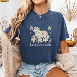May include: A denim blue short-sleeve t-shirt featuring a cream-colored graphic of a large sheep with three small lambs, surrounded by flowers and stars. The text "Raising Little Lambs" is printed in a serif font below the sheep illustration.