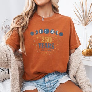 Op de afbeelding: Verbrand oranje t-shirt met een afbeelding ter viering van "250 JAAR" met maanfase-illustraties en de jaren 1776-2026. Het shirt is effen van kleur en heeft een ronde hals.