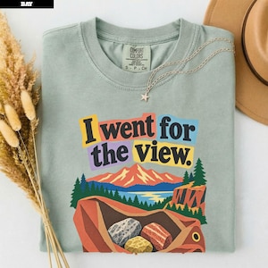 以下が含まれることがあります： I went for the viewというテキストがプリントされたセージグリーンの半袖Tシャツ。グラフィックには山脈、湖、松の木、3つの石が入った茶色のバッグが描かれています。Tシャツの内側の襟元にはComfort Colorsのラベルが付いています。