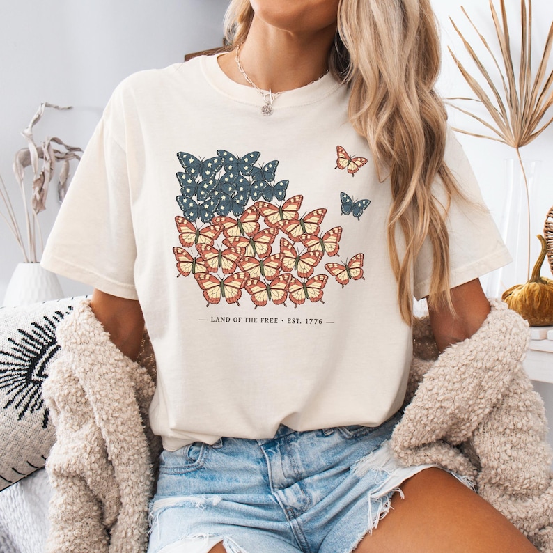 以下が含まれることがあります： 赤、白、青の蝶のデザインが施されたクリーム色のTシャツ。旗に似ています。「LAND OF THE FREE - EST. 1776」の文字が下にプリントされています。Tシャツは半袖です。