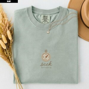 May include: A sage green crewneck t-shirt featuring an embroidered compass graphic above the word seek and the text Matthew 6:33. The shirt has a Comfort Colors label at the inner neckline and is accented with a delicate silver star pendant necklace.