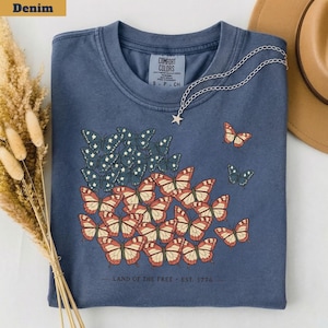 以下が含まれることがあります： デニムブルーのTシャツ。赤、白、青の蝶のデザインで、旗を連想させる。Tシャツには「LAND OF THE FREE - EST. 1776」の文字。星のペンダントが付いたシルバーのネックレスがTシャツにかけられている。
