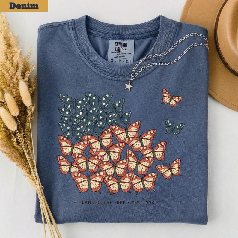 以下が含まれることがあります： デニムブルーのTシャツ。赤、白、青の蝶のデザインで、旗を連想させる。Tシャツには「LAND OF THE FREE - EST. 1776」の文字。星のペンダントが付いたシルバーのネックレスがTシャツにかけられている。