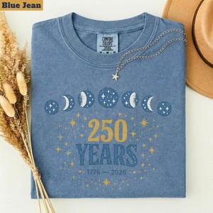 Op de afbeelding: Een blauw spijkerbroekkleurig T-shirt met een ronde hals. Het shirt heeft een ontwerp van maanfasen, sterren en de tekst "250 YEARS 1776-2026" in goud. Een zilveren ketting met een sterhanger ligt op het shirt.