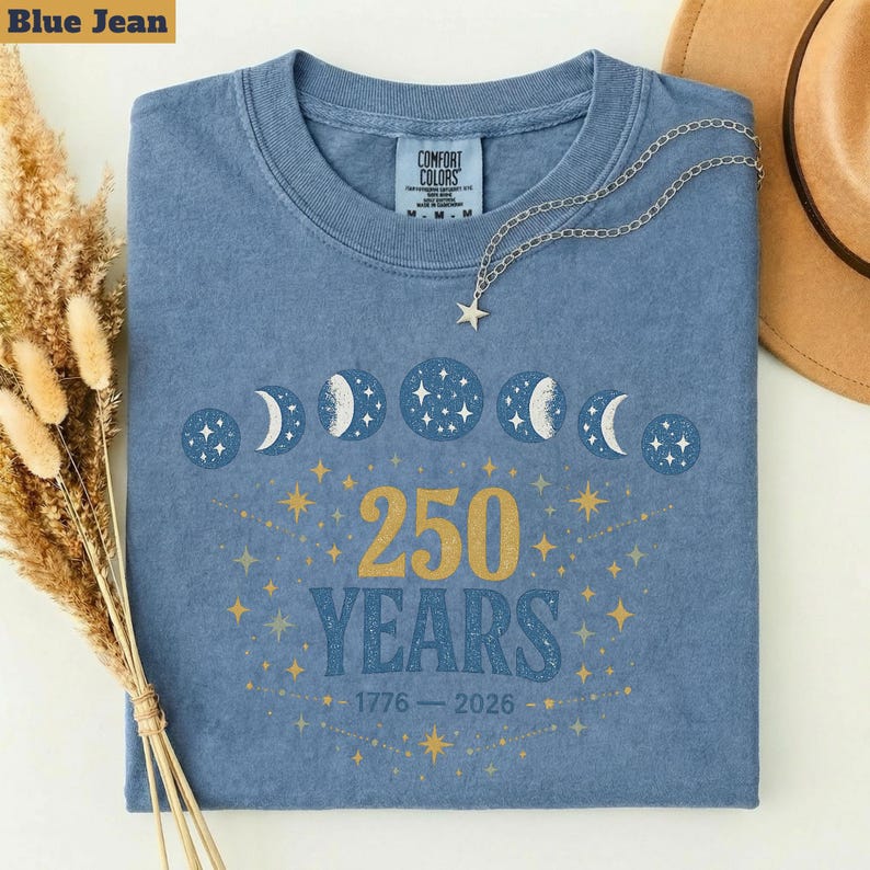 Op de afbeelding: Een blauw spijkerbroekkleurig T-shirt met een ronde hals. Het shirt heeft een ontwerp van maanfasen, sterren en de tekst "250 YEARS 1776-2026" in goud. Een zilveren ketting met een sterhanger ligt op het shirt.