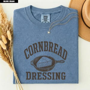 以下が含まれることがあります： クルーネックのジーンズブルーの半袖Tシャツ。フロントには、鋳鉄製のフライパンに入ったコーンブレッドのカットのダークブラウンのグラフィックが描かれており、そのイラストの上下に「CORNBREAD DRESSING」という文字がアーチ状に配置されています。