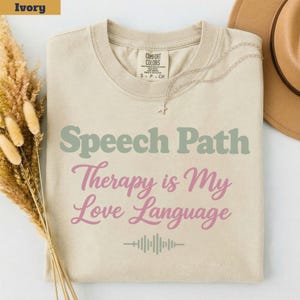 May include: An ivory crewneck t-shirt featuring the sage green text Speech Path and pink text Therapy is My Love Language, with a sage green sound wave graphic below. The shirt has a Comfort Colors brand label at the inner neckline.