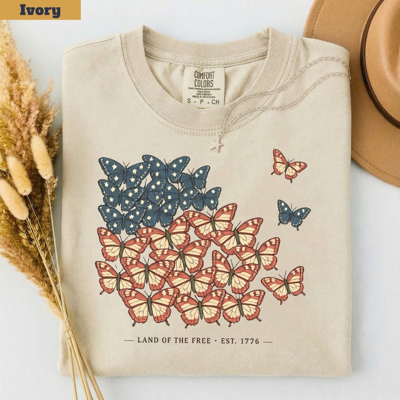以下が含まれることがあります： アイボリー色のTシャツ。アメリカ国旗を模した蝶のデザインが特徴です。白い星が付いた青い蝶と、赤とクリーム色の蝶が描かれています。「LAND OF THE FREE. EST. 1776」の文字がデザインの下にプリントされています。