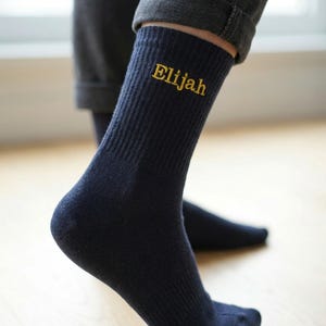 May include: A pair of navy blue ribbed crew socks featuring the name Elijah embroidered in yellow thread on the outer side of the ankle. The socks have a classic knit texture and reach mid-calf, providing a simple and functional design for daily wear.