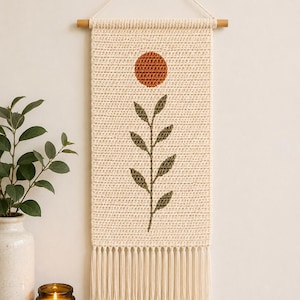 May include: Cream-colored, crocheted wall hanging with a wooden dowel and fringe. The design features a rust-colored sun and a green leaf stem. The wall decor is suspended by a white cord and a wooden bead.