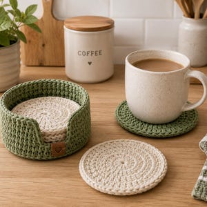 May include: A set of handmade crochet coasters in a holder. The coasters are off-white and the holder is olive green. A mug of coffee sits on a green coaster. A jar with the word "COFFEE" is in the background.
