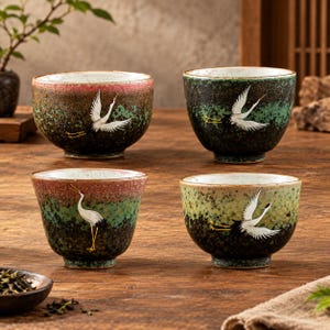 May include: Four ceramic tea cups with a speckled, multi-coloured glaze featuring hand-painted white cranes. Each cup displays a unique gradient of pink, green, and dark brown tones with a textured finish, showcasing flying or standing crane motifs.