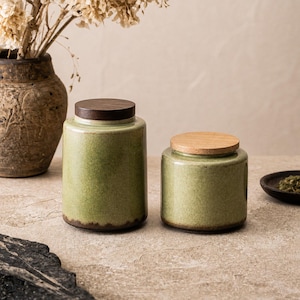Könnte beinhalten: Zwei grüne Keramikgläser mit Holzdeckeln. Die Gläser haben eine gesprenkelte Oberfläche und einen dunkelbraunen Boden. Ein Glas ist höher als das andere. Die Deckel sind hellbraun. Die Gläser stehen auf einer neutral getönten Oberfläche.