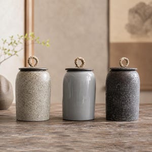 Può includere: Tre vasi in ceramica con coperchi. I vasi sono in tonalità di grigio e beige, con finitura strutturata o lucida. Ogni vaso ha un coperchio scuro e un manico in corda intrecciata. I vasi sono esposti su una superficie di legno.