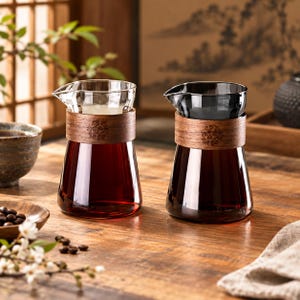 Op de afbeelding: Twee glazen kannen met een taps toelopende vorm en een brede houten band rond de hals. De linkerkan is van helder glas, terwijl de rechterkan van donkergrijs getint glas is. Beide hebben een kleine schenktuit en een florale houtsnijwerk.